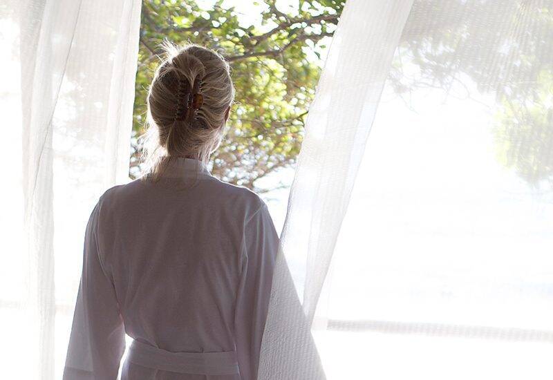 Woman wearing a white bath robe standing in front of a window with sheers.